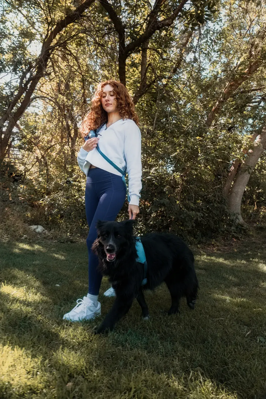 Woman with a Coast crossbody bag standing beside a black dog in a Coast harness in a sunny yard