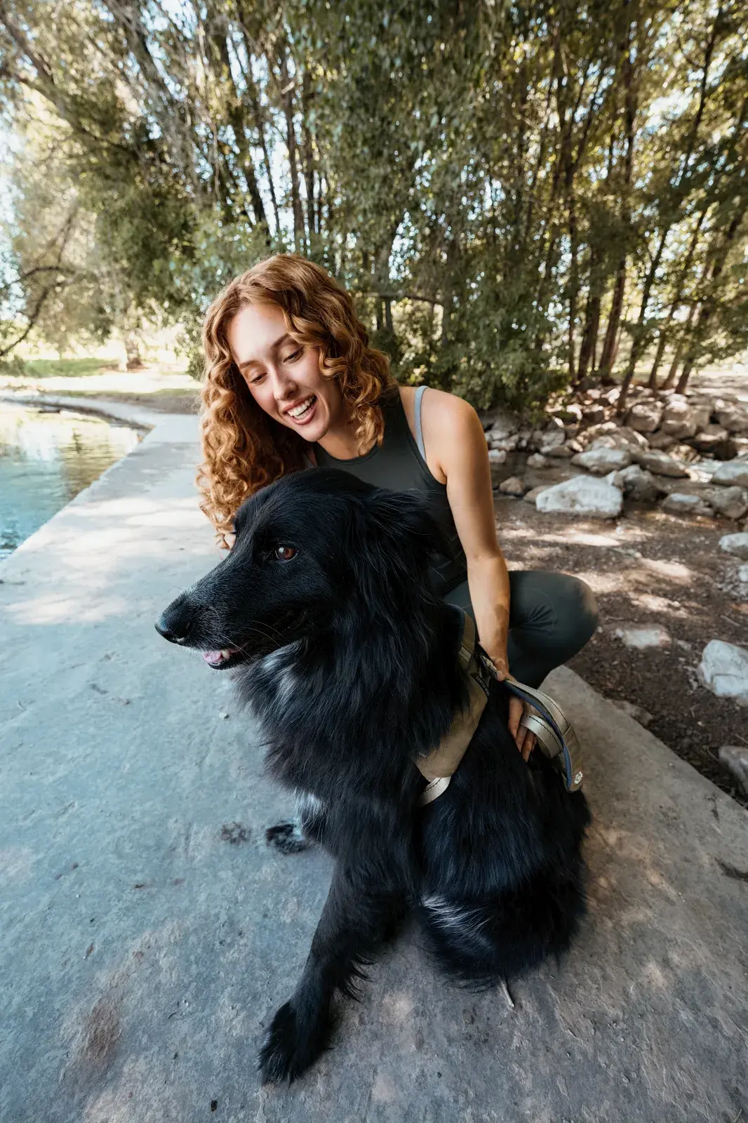 Woman crouching beside a fluffy black dog in a Grove harness near the water